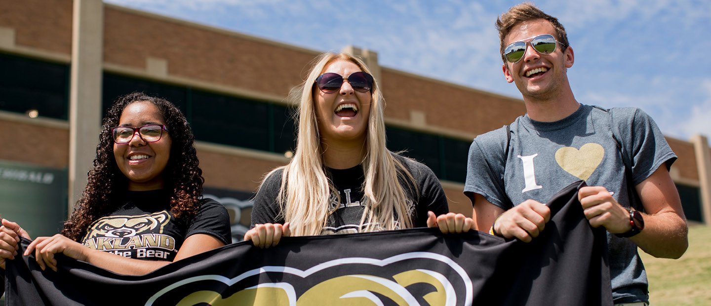 Three O U students holding a black flag with the gold Grizz head mascot on it.