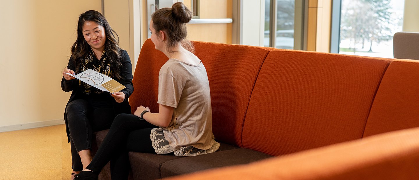 An adviser and a student seated on a couch, reviewing a brochure.