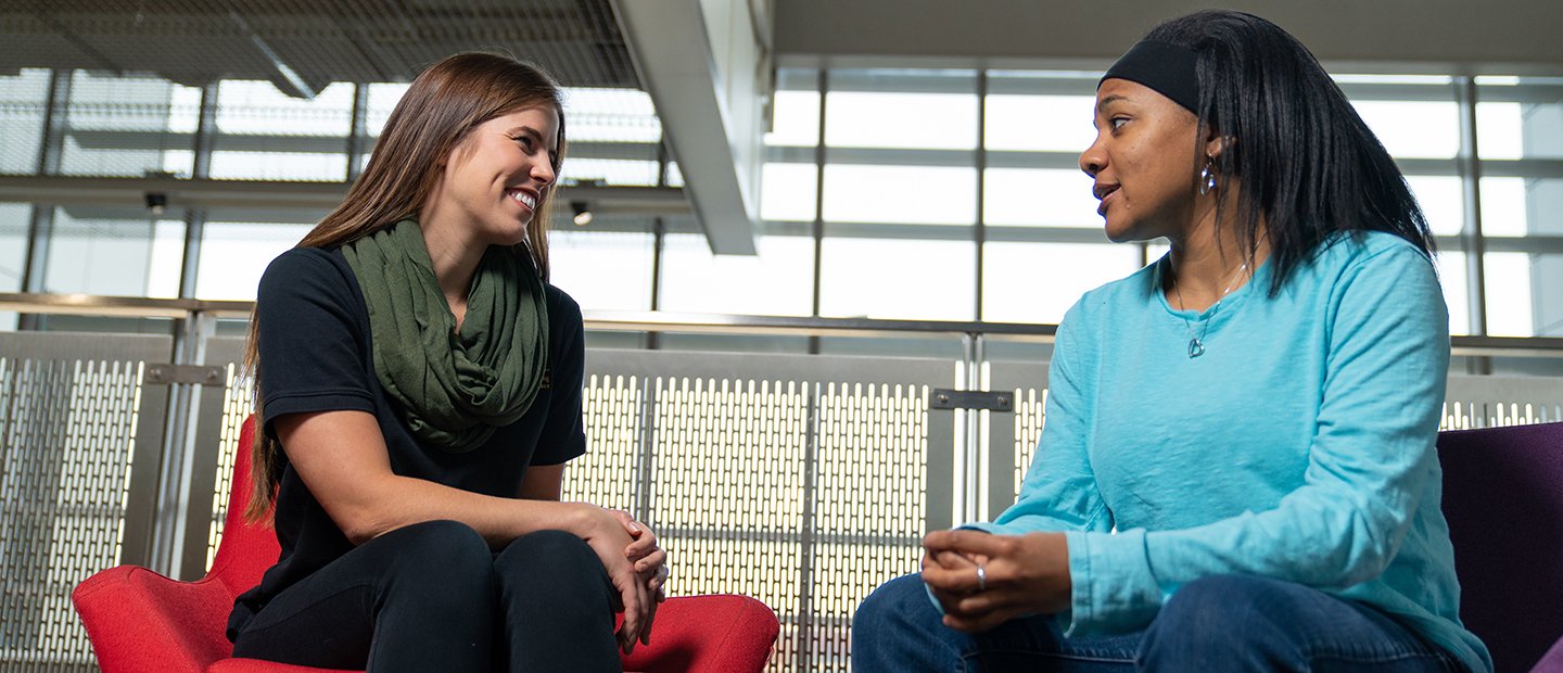 Two students seated in chairs, talking to each other.