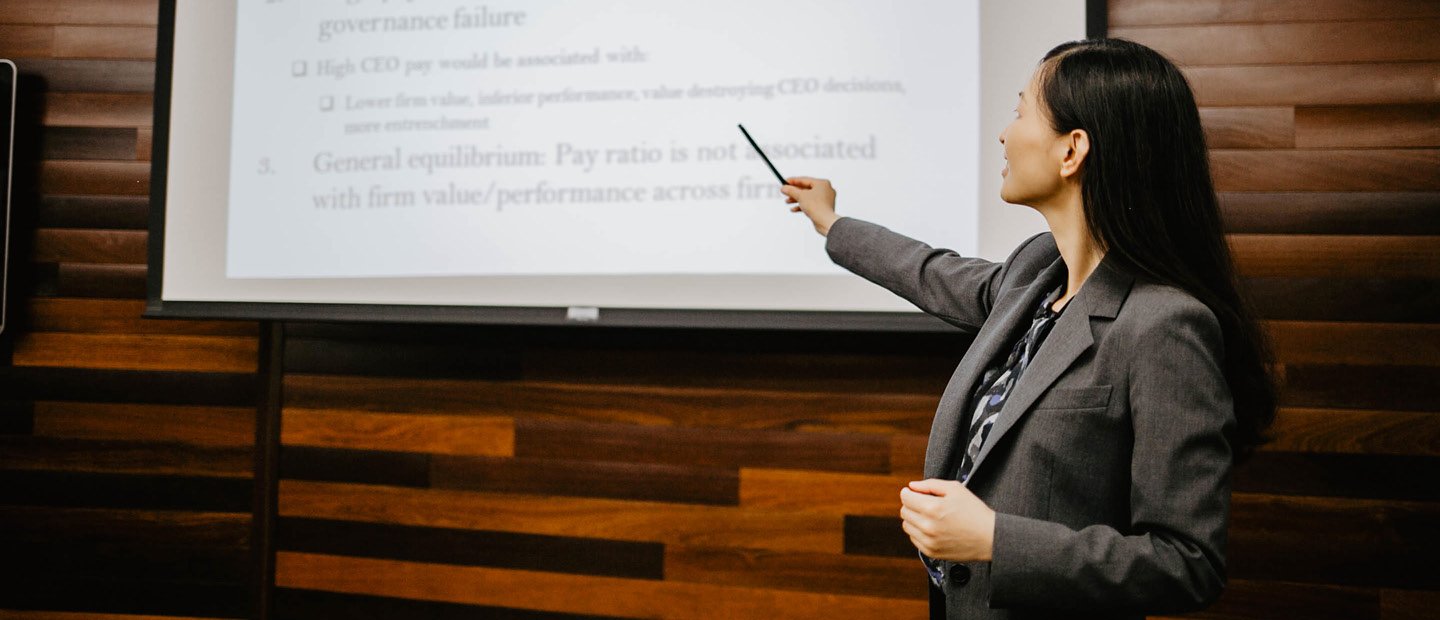 A woman in a suit standing in front of a projector screen, pointing at a line of text on it.