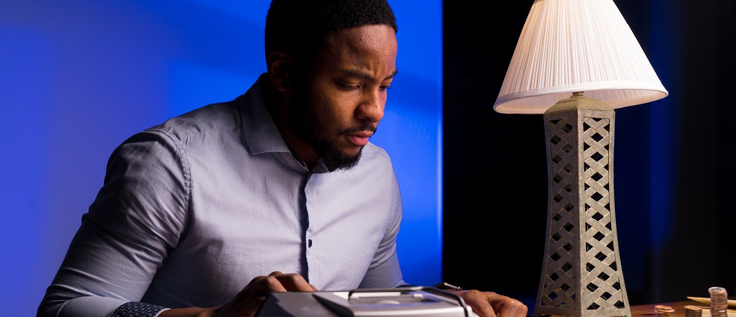 A man seated at a desk with a lamp, using a large calculator.