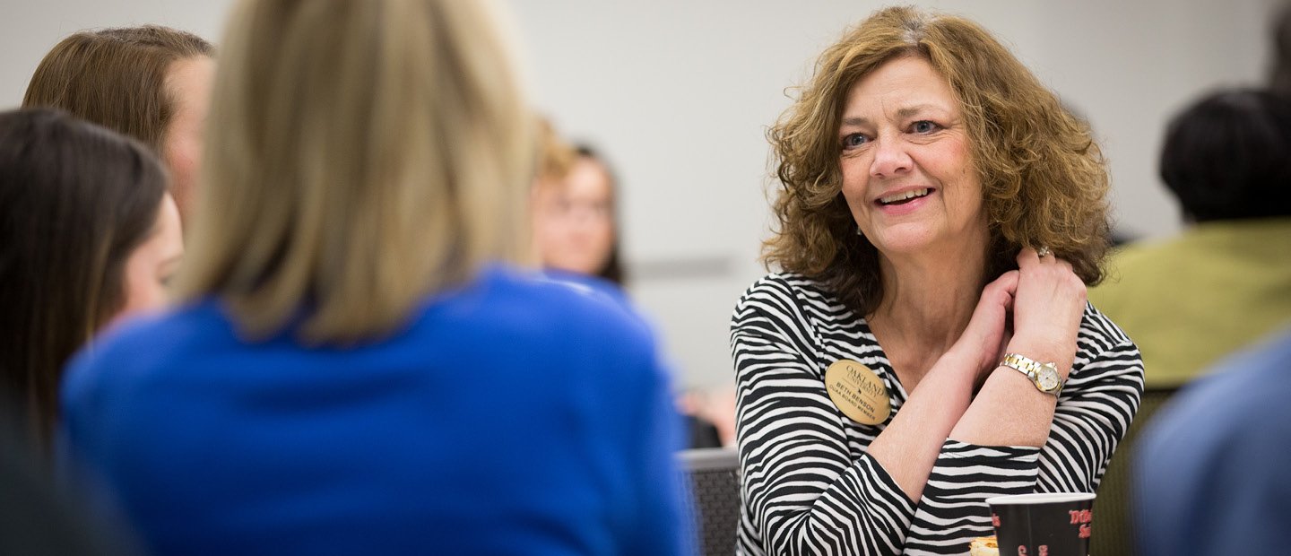 A woman wearing a name tag, seated, talking to three other women.