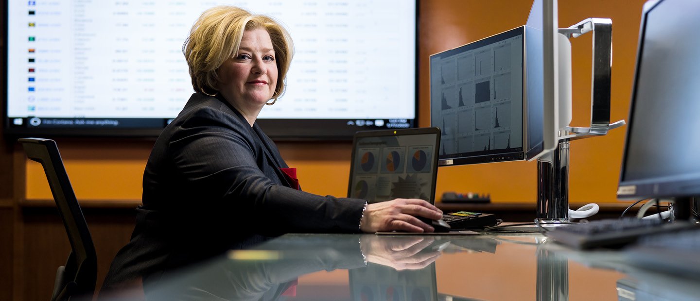 A woman seated a desk with multiple computer screens and a projector behind her, looking at the camera.