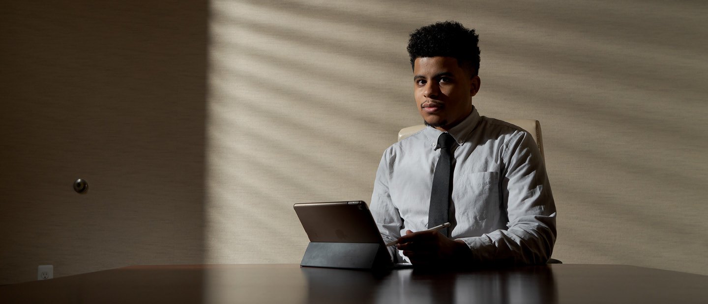 A young man seated a large table with a tablet in front of him, looking at the camera.