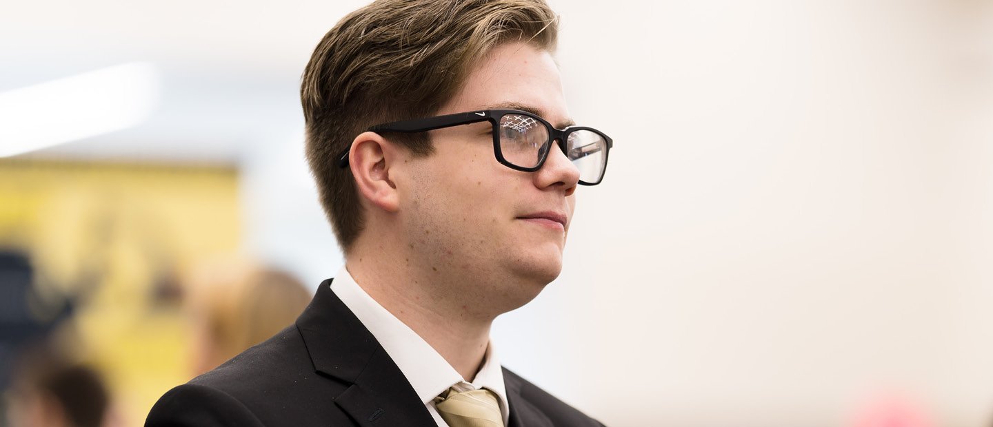 A profile photo of a young man in glasses and a black suit jacket with a gold tie.