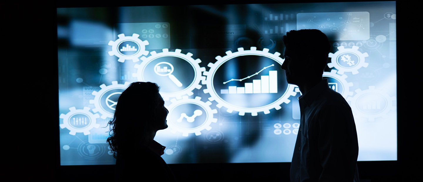 Silhouettes of two people in front of a blue screen displaying white gear and chart graphics.