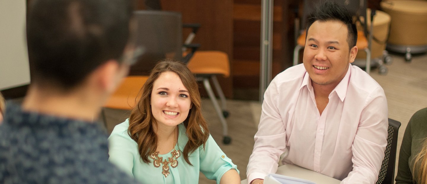 A man and woman seated at a desk, looking up and smiling at a man standing in front of them.