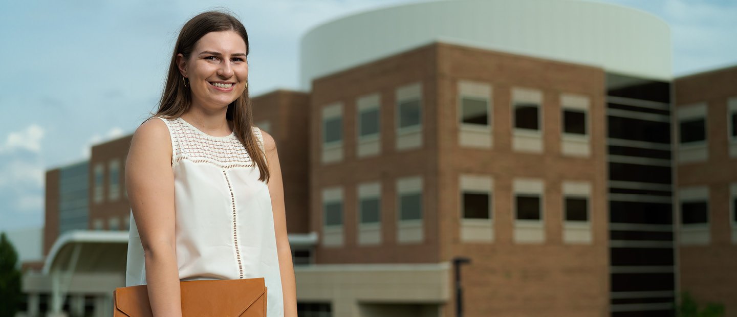 A woman holding a brown folder standing in front of a brown building at Oakland University.