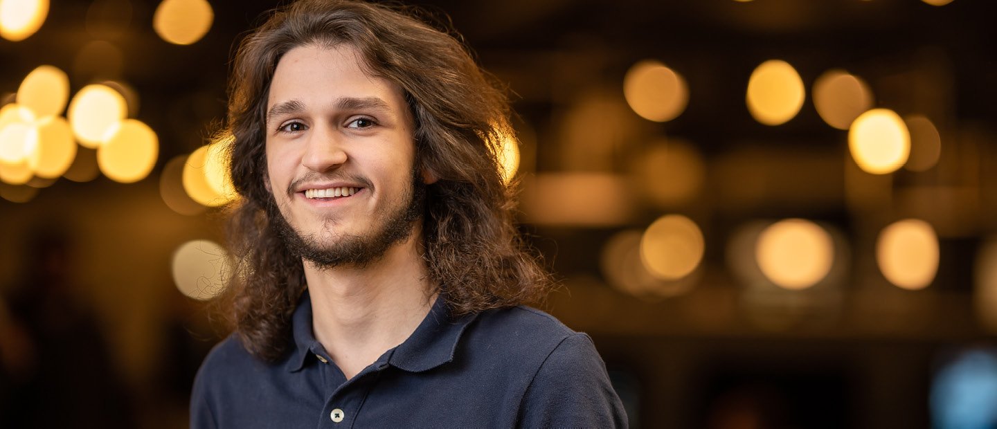 A young man smiling at the camera with blurred yellow lights behind him.