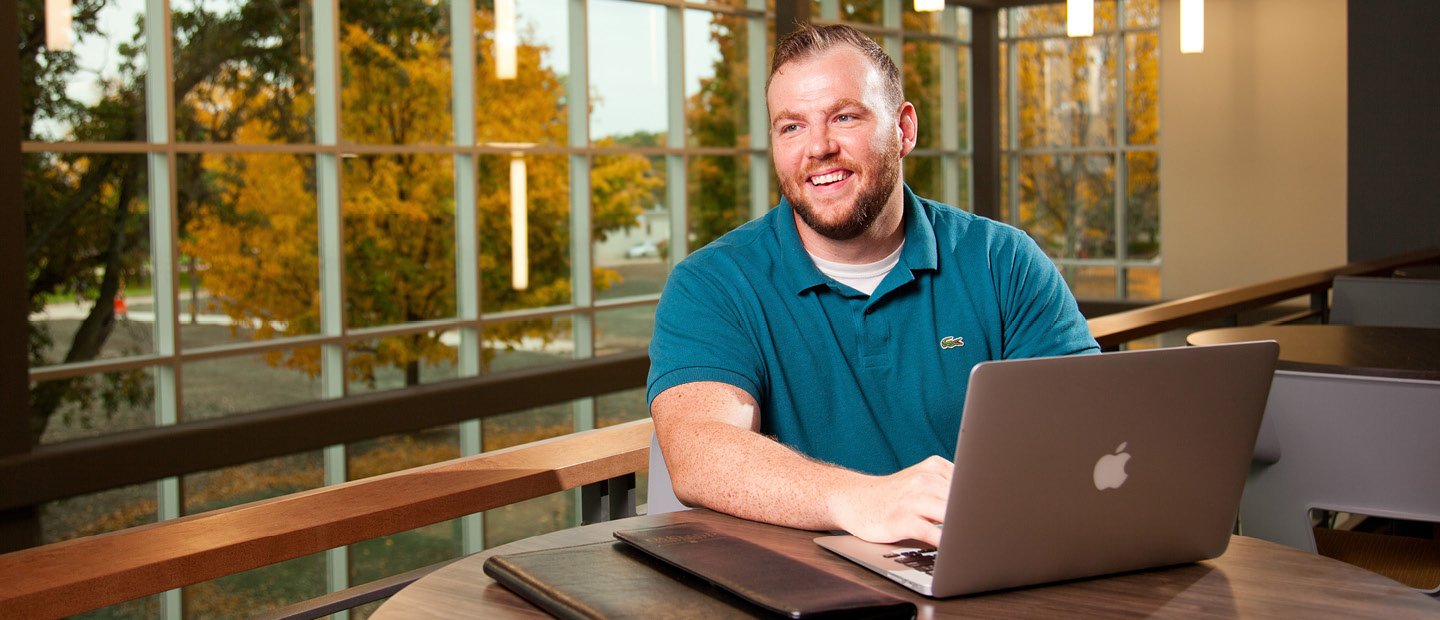 A man seated at a table with a laptop and a view of trees with yellow leaves through windows behind him.