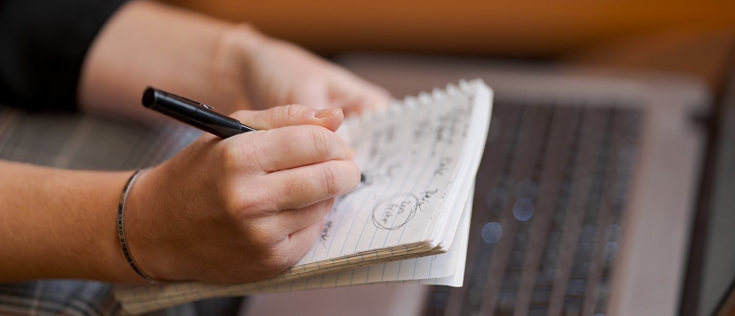 A person's hands writing on a notepad, held over an open laptop.