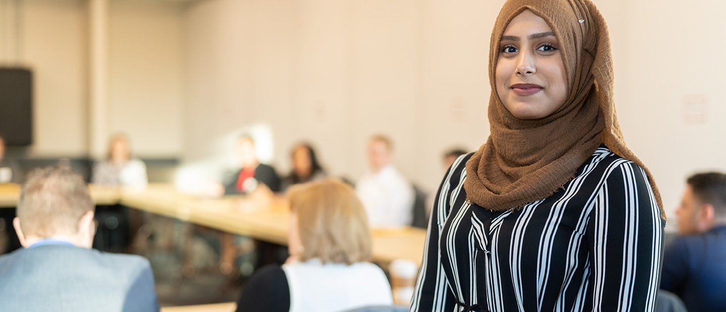 A woman smiling at the camera, standing at the back of a classroom full of people.