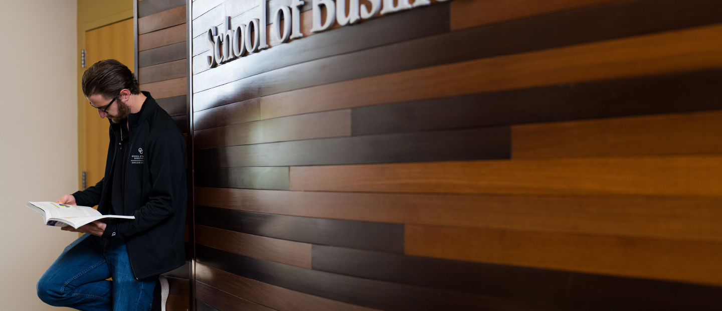 A man reading a book, leaning against a wall with the words School of Business Administration behind him.