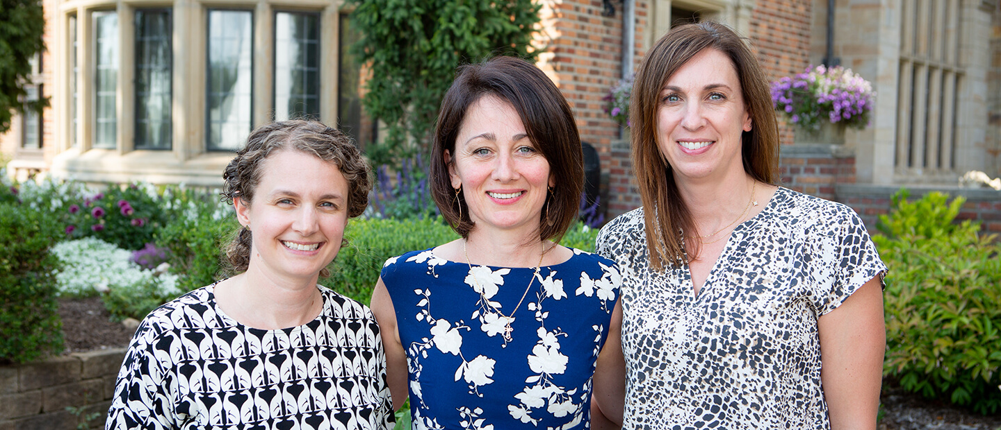 Three women, smiling, standing together in front of garden