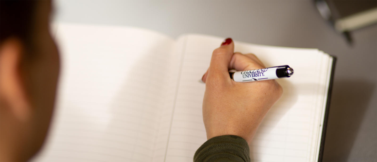 A person's hand holding an Oakland University pen over an empty notebook.