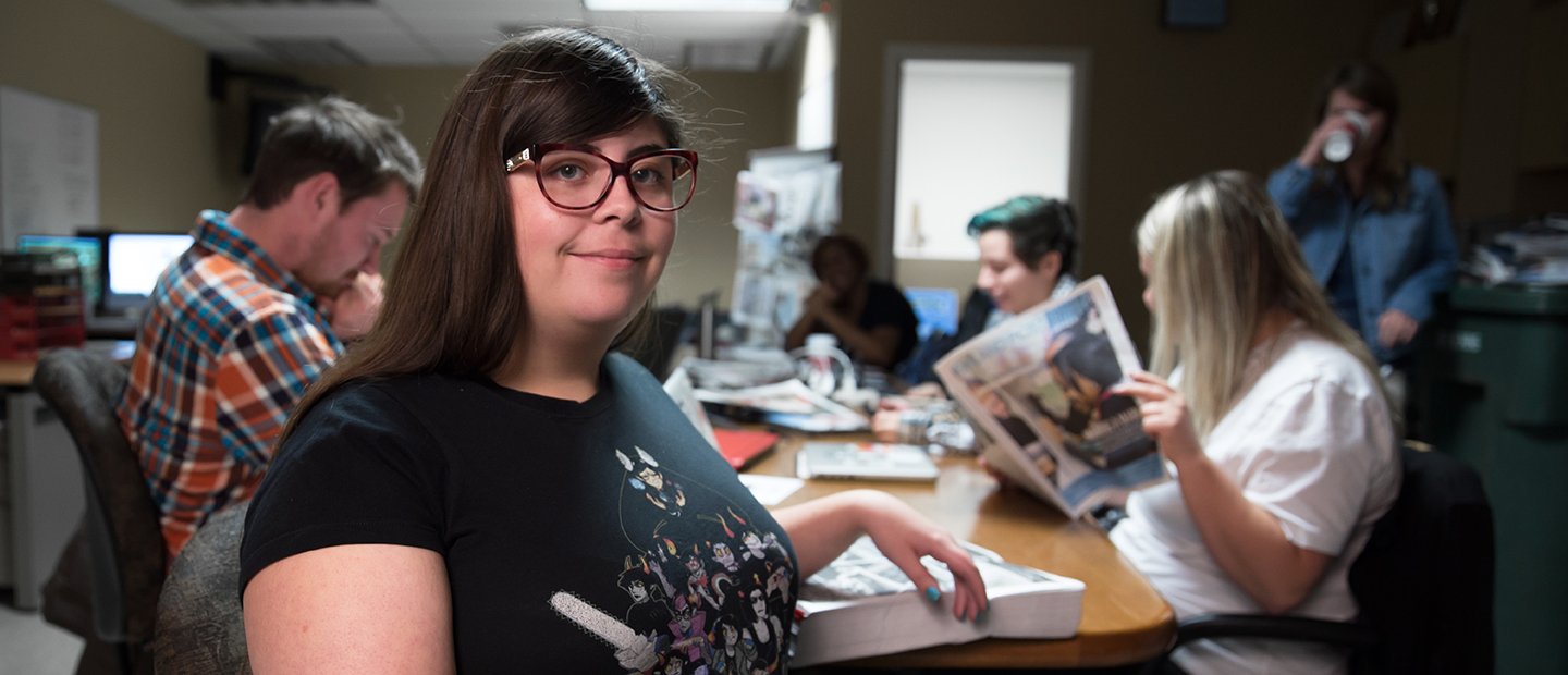 A woman faces the camera smiling. Her classmates are reading books and newspapers in the background