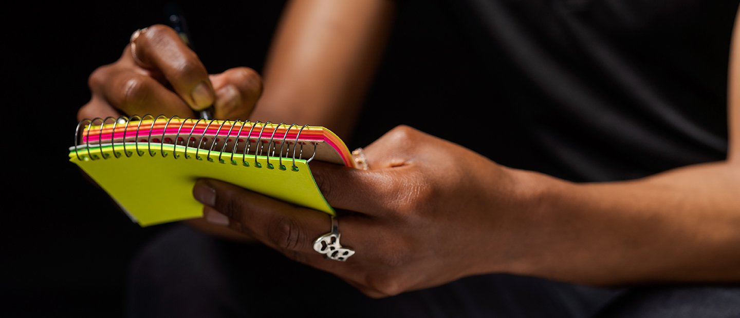 A person writing in a small notepad with brightly colored paper.