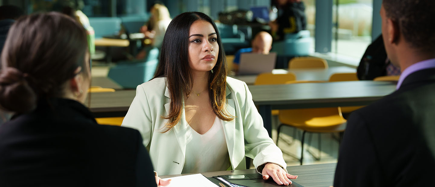 Student sitting at a table having a meeting with a man and a woman with a portfolio in front of her on the table.