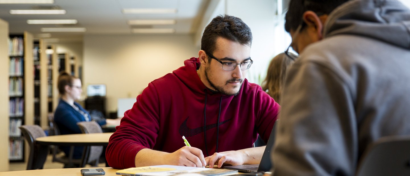 A male student looking at his laptop is writing notes on a pad of paper