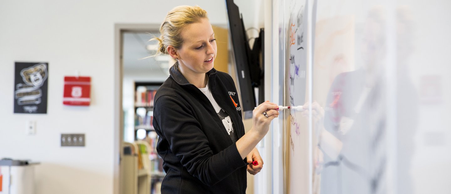 A woman in front of a classroom writes on a whiteboard.