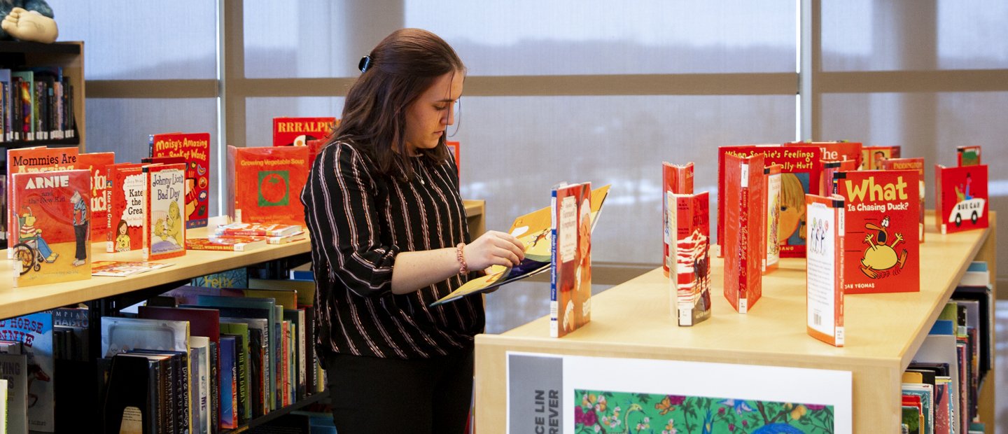 A woman standing by a bookshelf reviews a children picture book.