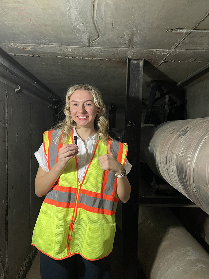 Mallory Waligora holds up a microphone as she wears a bright yellow vest in a tunnel.