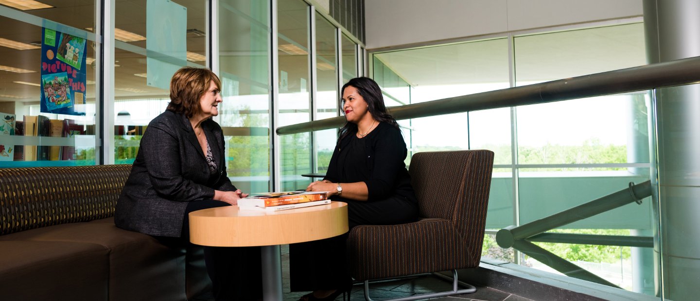 two women seated at a round table with books on it, windows behind them