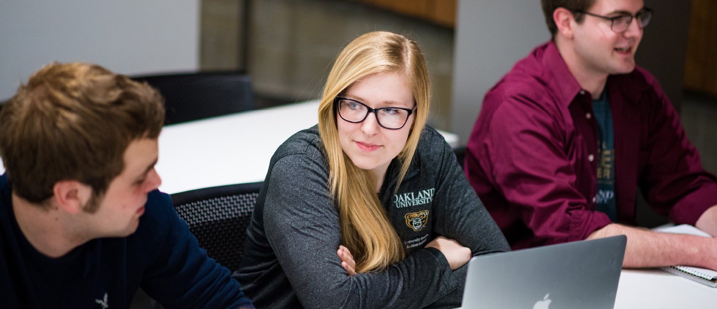 female student wearing a grey Oakland University shirt with an open laptop in front of her, male student on her right and left