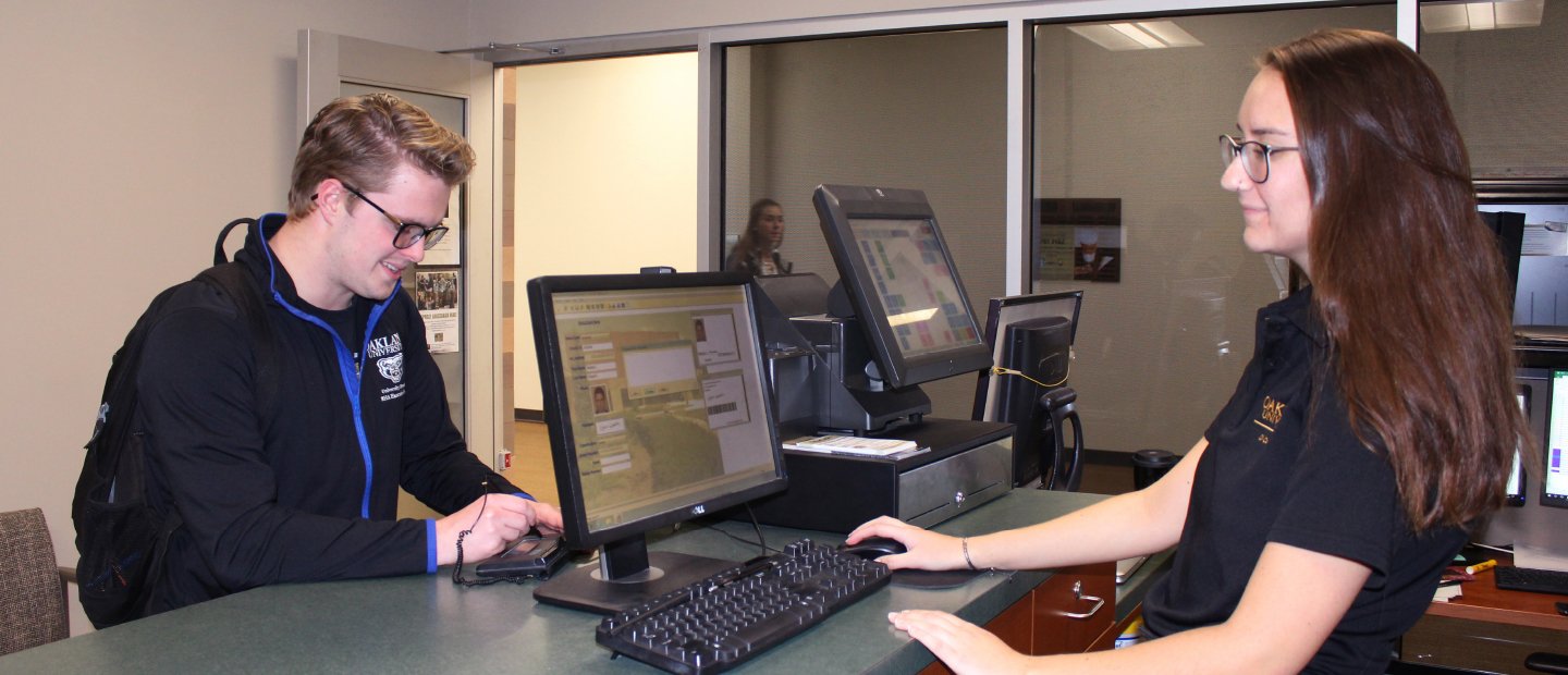 young woman standing behind a desk with a computer, assisting a young man on the other side of the counter