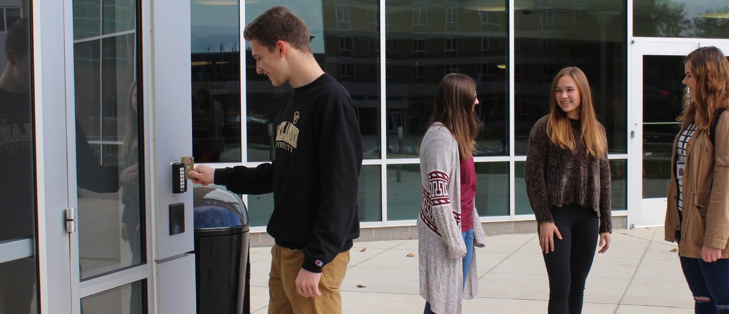 young man swiping an ID card through a reader on a wall outside, three young women in the background