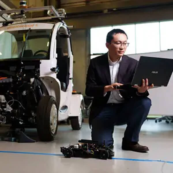 Man kneels in front of a prototype vehicle. He is looking at a laptop that he holds in his hands.