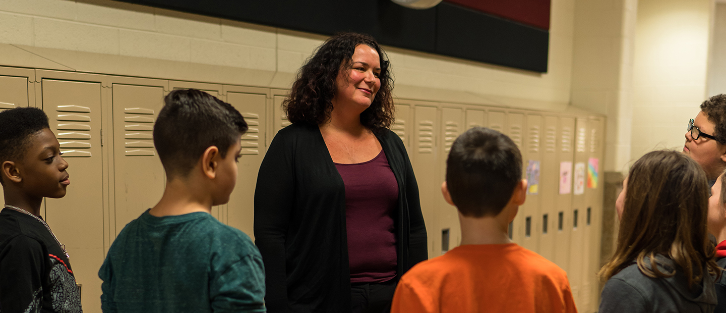 Educational Leadership Banner A woman standing in a hallway with lockers behind her and young students in front of her.