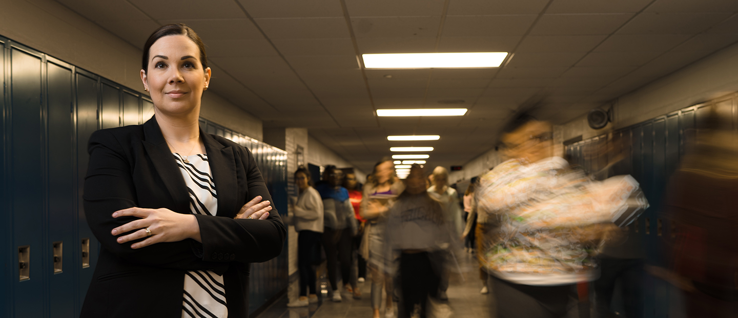 Graduate Programs Banner A woman standing with her arms folded across her chest, in a hallway of a school with blue lockers along the wall and people walking behind her.
