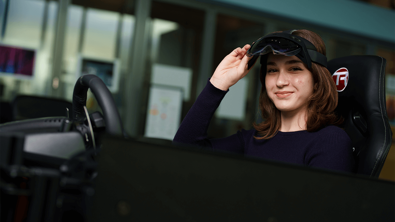 A woman wearing safety goggle sits behind a steering wheel.