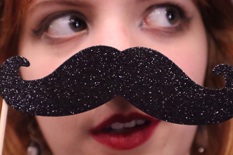 A woman holds a paper moustache in front of her face.