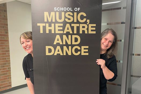 Two women stand next to a sign that says Music, Theatre and Dance