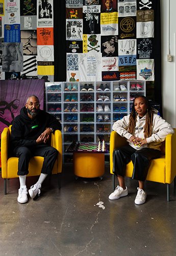 Two people sitting in chairs with a collection of shirts and shoes behind them