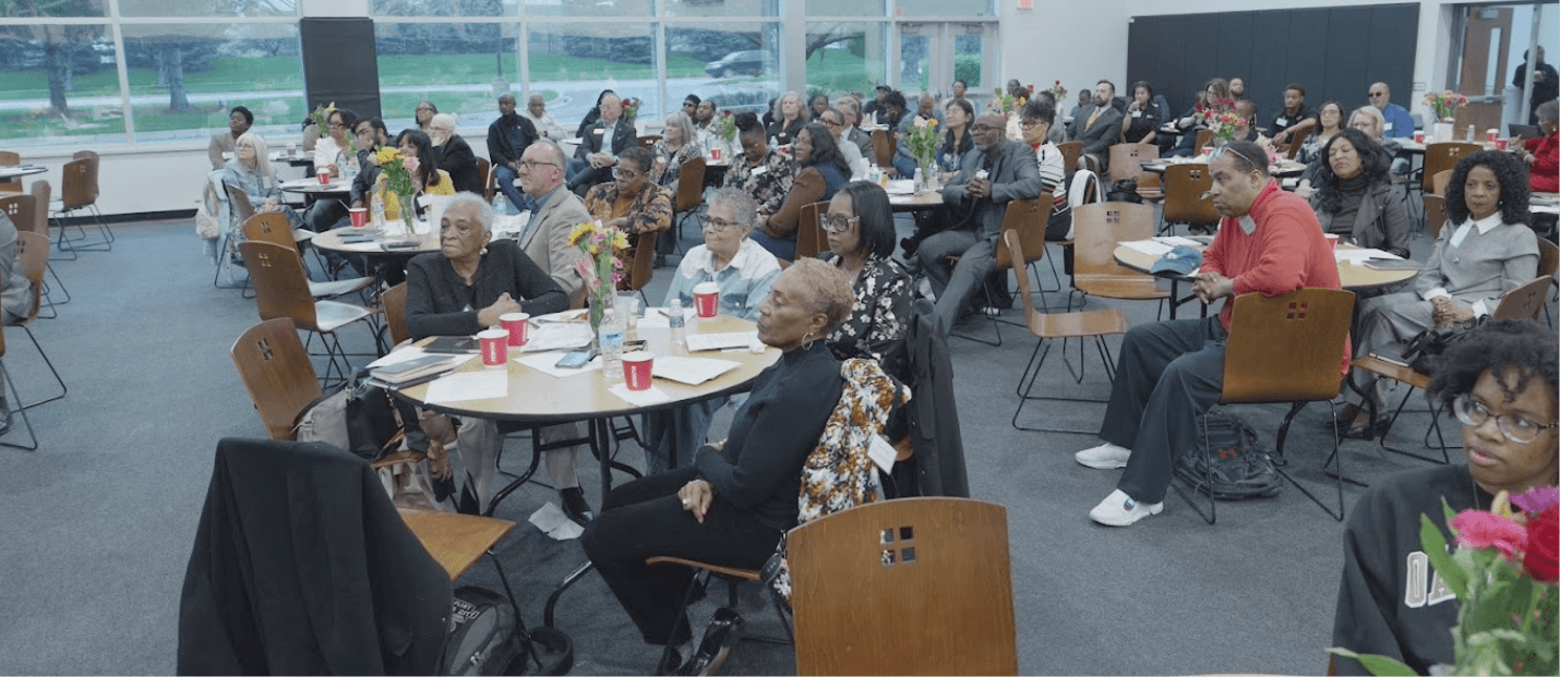 People sitting at tables listening to a speaker in a room with large windows.