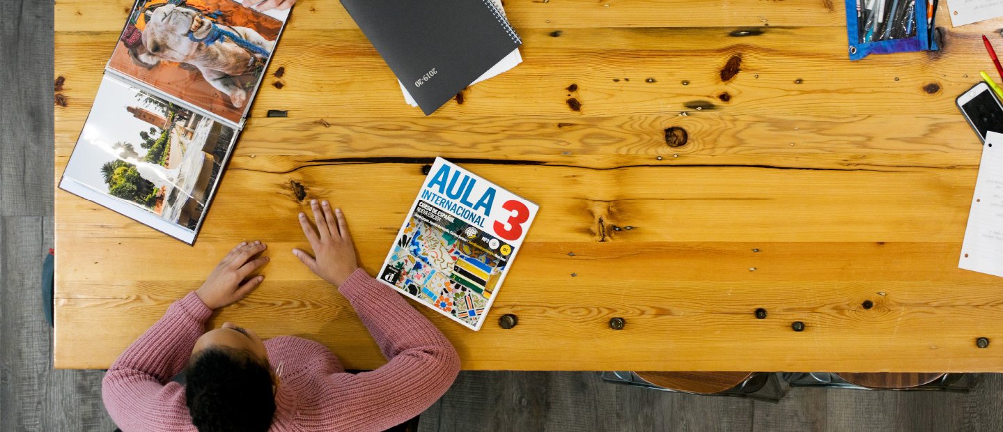 An aerial photo of a woman seated at a wooden table with books and notebooks in front of her.