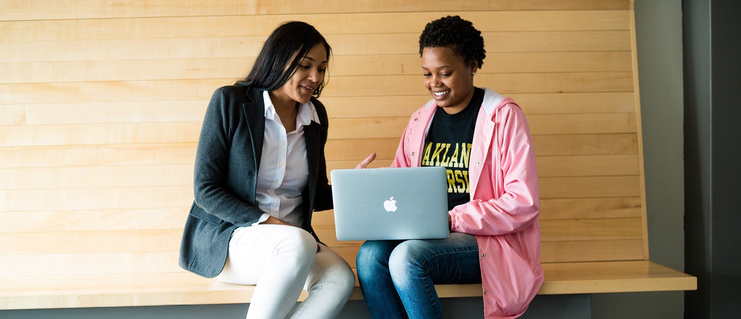 two young women seated on a wooden bench, looking at a laptop screen.