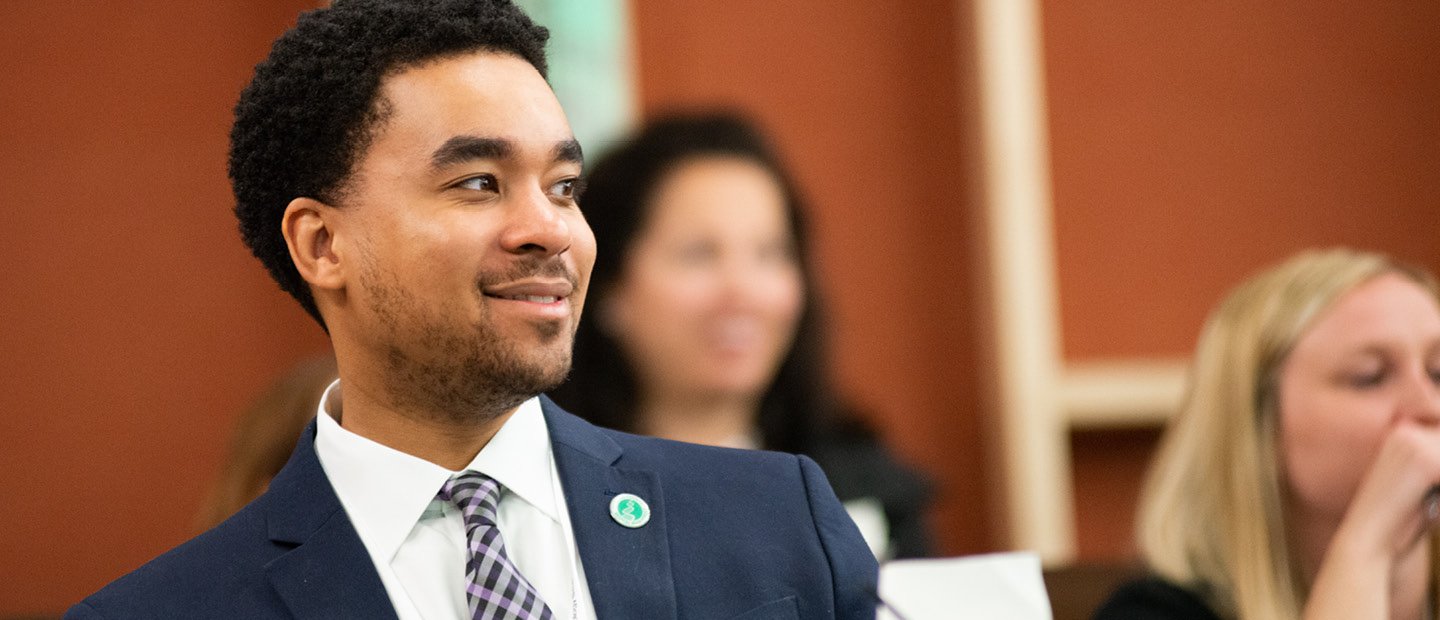 young man in a suit, seated in a room full of people.