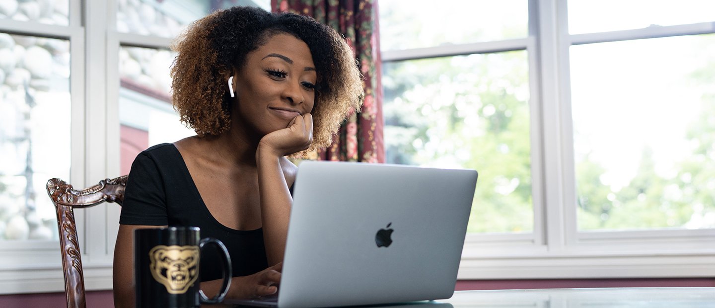 A female student seated at a table with an open laptop and a coffee mug with a gold Oakland University Grizz bear image on it.