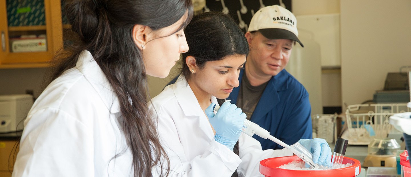 A woman holding a vial in a lab while two people observe.