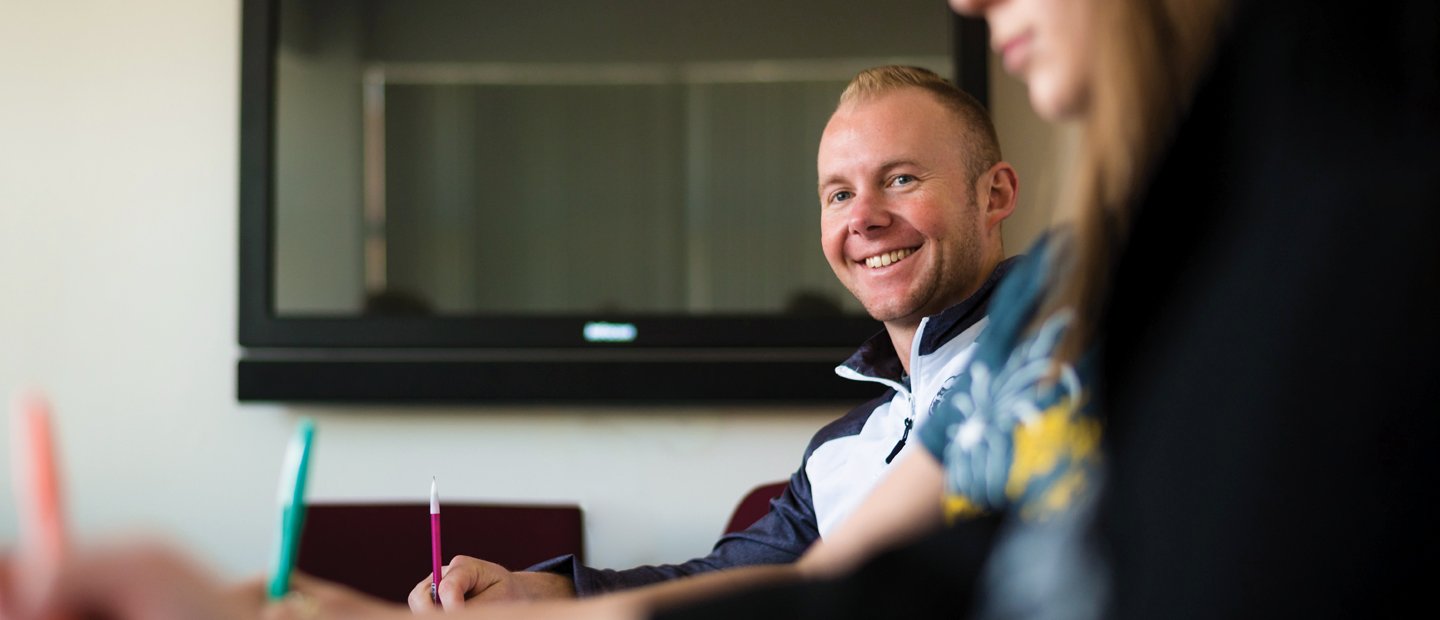 A man at a table with other students, smiling at the camera.