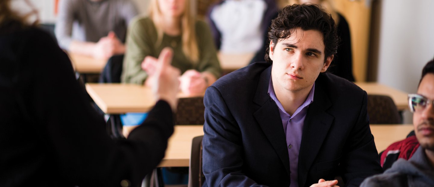 young man in a classroom full of students, watching someone at the front of the room