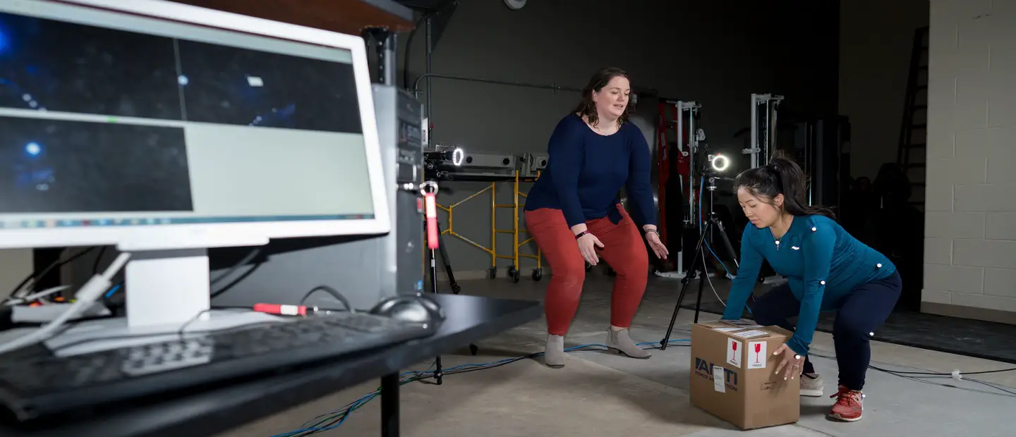 A woman showing another woman how to safely lift a heavy box in a workplace.