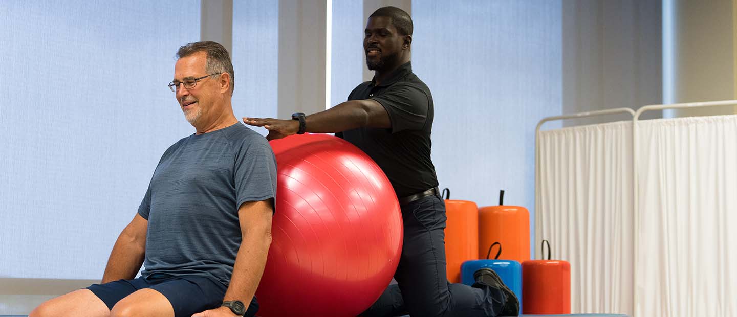 A male physical therapist helping a man with an exercise ball on a table in a fitness studio.