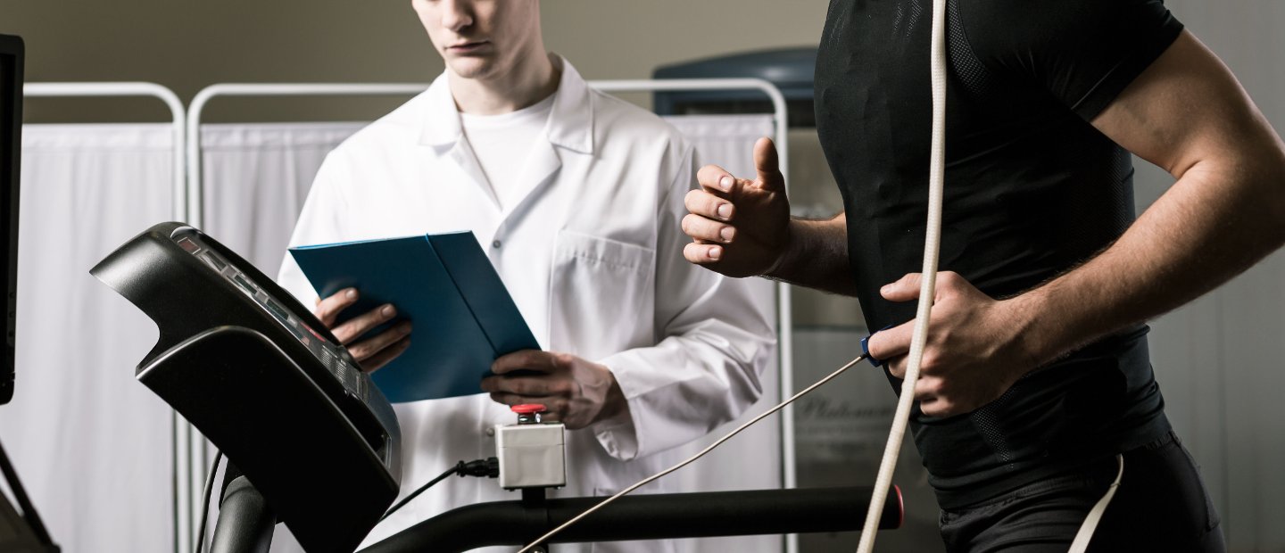 A patient running on a treadmill while a doctor holding a chart observes.