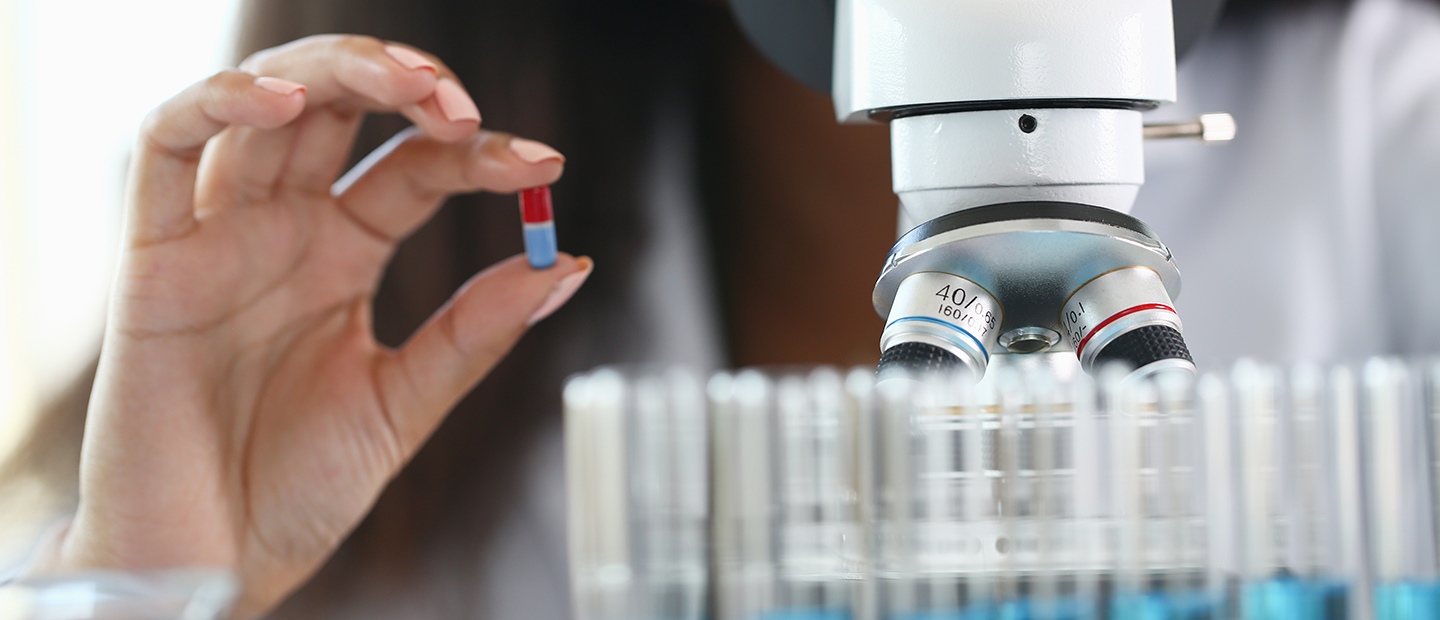 A close up image of a hand holding a pill between thumb and index finger, next to a microscope.