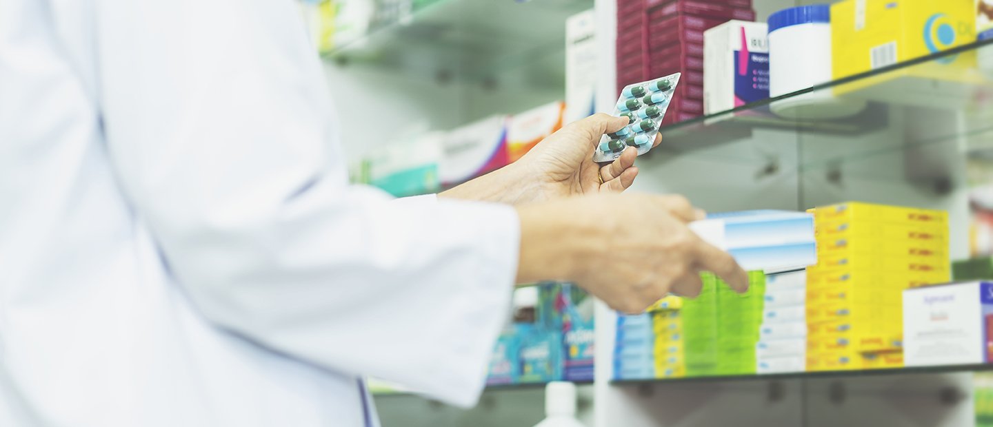 Person in a white lab coat holding a pack of pills in front of a shelf of medicine.